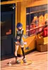 A figurine of a young woman standing in front of a building with yellow wooden walls and a dark blue door. Produktbild-Vorschau
