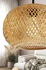 A close-up of a woven bamboo pendant light fixture in a living room setting, with blurred furniture and plants in the background. Náhled obrázku produktu
