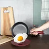 A hand is holding a cast iron pan with a fried egg over a red plate, with cutting boards and kitchen towels in the background. Náhľad obrázku produktu