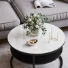 A round coffee table with a white top and black base, decorated with a vase of flowers, a small bowl of candies, and a candle holder, placed in a living room setting next to a gray sofa. Produktbild-Vorschau