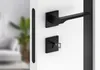 A modern black door handle set with a keyhole lock, mounted on a white door. The background is blurred, showing a hint of a living room with a plant. Produktbild-Vorschau