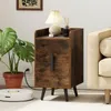 A rustic brown nightstand with two drawers and black metal legs is placed next to a beige sofa. A Monstera plant in a terracotta pot and a white robot vacuum cleaner are also visible. Podgląd zdjęcia produktu