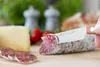 A close-up of a knife slicing a salami, with cheese, tomatoes, and bread in the background. Voir les images du produit