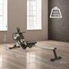 A rowing machine is shown in a room with windows and a brick wall. Dimensions are indicated. Produktbild-Vorschau