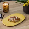 A waffle on a yellow plate on a wooden table, next to a lit candle and a vase of flowers. Náhľad obrázku produktu