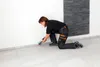 A person is kneeling and applying sealant to the base of a wall where it meets the floor. Produktbild-Vorschau