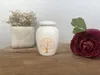 A small white urn with a gold tree design sits on a wooden surface, next to a potted plant and a dark red flower. Náhled obrázku produktu