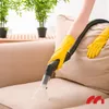 A person wearing yellow gloves is using a wet/dry vacuum cleaner to clean a beige sofa. There are plants in the background. Produktbild-Vorschau
