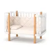 A white baby crib with wooden accents and a cream-colored mattress and bumper pad. Produktbild-Vorschau