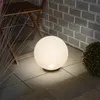 A glowing sphere-shaped lamp sits on a tiled patio next to a wooden chair and some dry grass. Produktbild-Vorschau