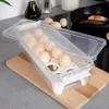 A clear plastic egg container filled with eggs sits on a wooden cutting board on a kitchen counter. In the background, a frying pan with a fried egg is visible. Podgląd zdjęcia produktu
