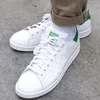 A close-up shot of a pair of white Adidas Stan Smith sneakers with green accents, worn with khaki pants and white socks, resting on a concrete surface. Náhled obrázku produktu