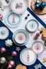 A collection of Christmas-themed tableware, including plates and bowls, arranged on a blue and white striped cloth, with festive decorations like ornaments and pinecones scattered around. Anteprima dell’immagine prodotto