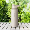 A beige water bottle sits on a white wooden table outdoors, with green foliage in the background. Produktbild-Vorschau