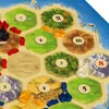 A close-up view of a board game, likely 'Settlers of Catan', showing hexagonal tiles with numbers and resource illustrations, along with game pieces. Náhled obrázku produktu