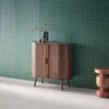 A wooden sideboard with fluted doors stands against a green tiled wall. A small copper stool is to the right. Produktbild-Vorschau