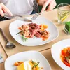 A person is eating a meal with steak, mushrooms, and vegetables on a white plate, with another plate of food and a glass of water in the background. Náhled obrázku produktu