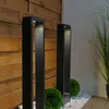 Two black bollard lights are placed in a garden setting, next to a wooden fence and some greenery. Anteprima dell’immagine prodotto