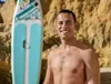 A man is smiling on a beach next to a paddleboard. Produktbild-Vorschau