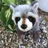 A plush raccoon toy is shown sitting on a bed of small stones, with green leaves in the background. Podgląd zdjęcia produktu
