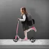 A young girl riding a pink scooter on a sidewalk in front of a gray wall. Náhľad obrázku produktu