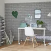 A home office or craft room setup with a desk, chair, storage unit, and decorative elements against a brick-patterned wall. Produktbild-Vorschau