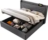 A grey upholstered storage bed with the mattress lifted to reveal storage compartments filled with pillows and folded items. A phone is charging on the headboard. Produktbild-Vorschau
