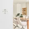 A white wall with a white smart home device mounted on it. The right side of the image shows a blurred interior of a dining room with a wooden table and chairs. Produktbild-Vorschau