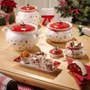 A collection of Christmas-themed ceramic dishes and containers arranged on a wooden table, with a decorated Christmas tree in the background. Náhled obrázku produktu