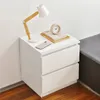 A white bedside table with a lamp, books, and a phone on top, placed next to a bed. Produktbild-Vorschau