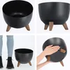 A collage of four images showing a black planter with wooden legs from different angles, including a person's legs and feet next to it for scale. Náhled obrázku produktu