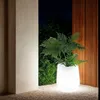 A glowing white planter with a green plant sits on gravel next to a textured wall and a slatted wooden screen at night. Náhled obrázku produktu