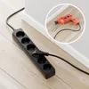 A black power strip with multiple outlets is plugged into the wall on a wooden floor. A circular inset shows a close-up of a red drill and screws. Produktbild-Vorschau