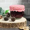A jar of jam with a red and white checkered lid, decorated with natural elements like pinecones and nuts, sits on a wooden slice. Produktbild-Vorschau
