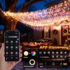 A hand holding a smartphone displaying a lighting control app, with a string of colorful icicle lights draped over a tree and house in the background, set in a snowy outdoor environment during the evening. Produktbild-Vorschau