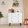 A white dresser with black knobs is shown in a room with plants and decorative items. Náhled obrázku produktu