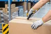 A person wearing gloves is using a utility knife to open a cardboard box in a warehouse. Anteprima dell’immagine prodotto