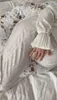 A person's hand rests on a cream-colored pregnancy pillow with a brown leaf pattern, placed on a white bed. Náhľad obrázku produktu