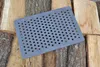 A perforated metal grate rests on a pile of cut firewood. Náhled obrázku produktu