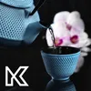 A light blue cast iron teapot pouring tea into a matching cup, with a pink and white orchid in the background. Náhled obrázku produktu