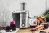 A juicer is shown on a wooden table with fresh ingredients and juice. Náhľad obrázku produktu