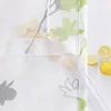 Close-up of a white fabric curtain with a floral pattern, featuring green and yellow flowers and gray leaves. A clear plastic tube is visible on the right side, and three lemons are placed on the white background. Produktbild-Vorschau