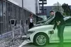 Two people are charging electric cars at charging stations outside a modern building. Produktbild-Vorschau