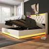 A modern bedroom with a white upholstered bed frame featuring LED lighting and a tufted headboard. The bed is shown with its storage compartment open, revealing a slatted mattress base. Produktbild-Vorschau