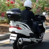 A person riding a white scooter on a road with greenery and flowers in the background. Anteprima dell’immagine prodotto