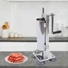 A stainless steel sausage stuffer machine is shown on a countertop next to a plate of sausages, with kitchen cabinets and shelves in the background. Náhled obrázku produktu