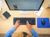 A person's hands are typing on a black keyboard, with a computer monitor, mouse, and notebook visible on a wooden desk. Produktbild-Vorschau