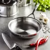 A stainless steel frying pan is placed on a countertop with other kitchen items like garlic, chili peppers, peppercorns, and a knife. Anteprima dell’immagine prodotto