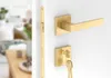 A close-up view of a golden door handle and lock set mounted on a white door, with a blurred interior scene visible through an open doorway in the background. Produktbild-Vorschau