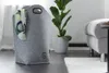 A grey felt laundry basket with a scarf draped over the side, placed on a dark wooden floor next to a potted plant and a sofa. Produktbild-Vorschau